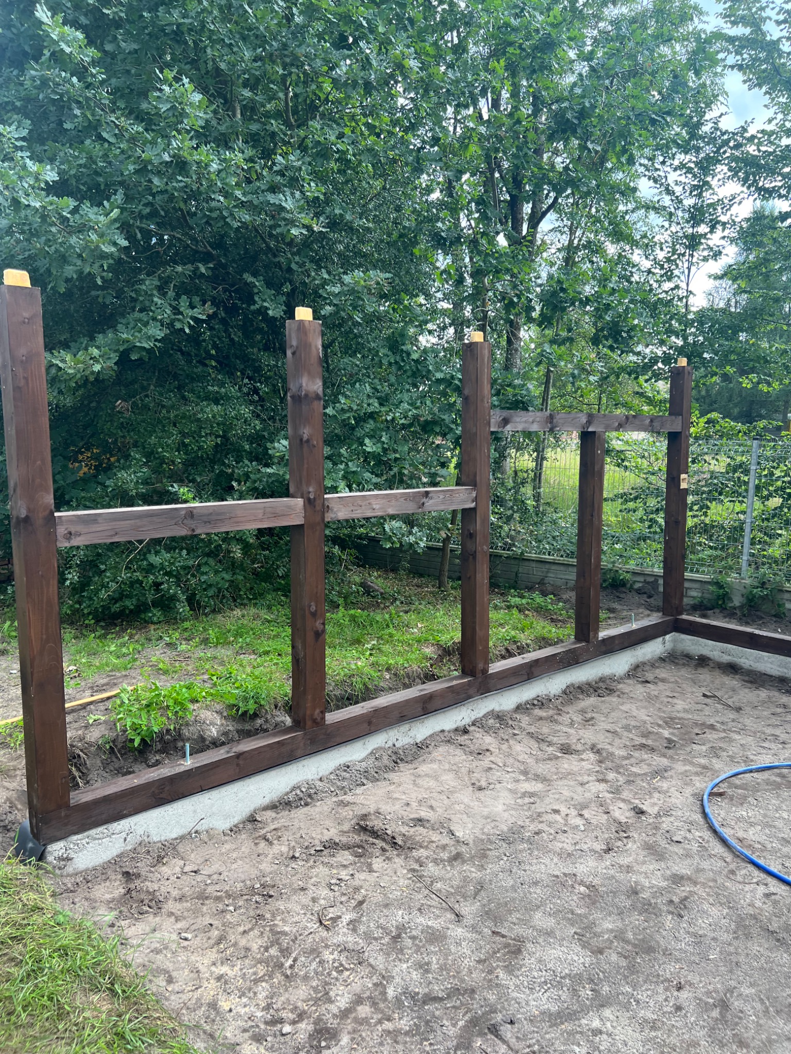 Wczesny etap budowy konstrukcji z ciemnego drewna, prawdopodobnie altany lub wiaty, na betonowej podstawie. W tle widoczne drzewa i krzewy.