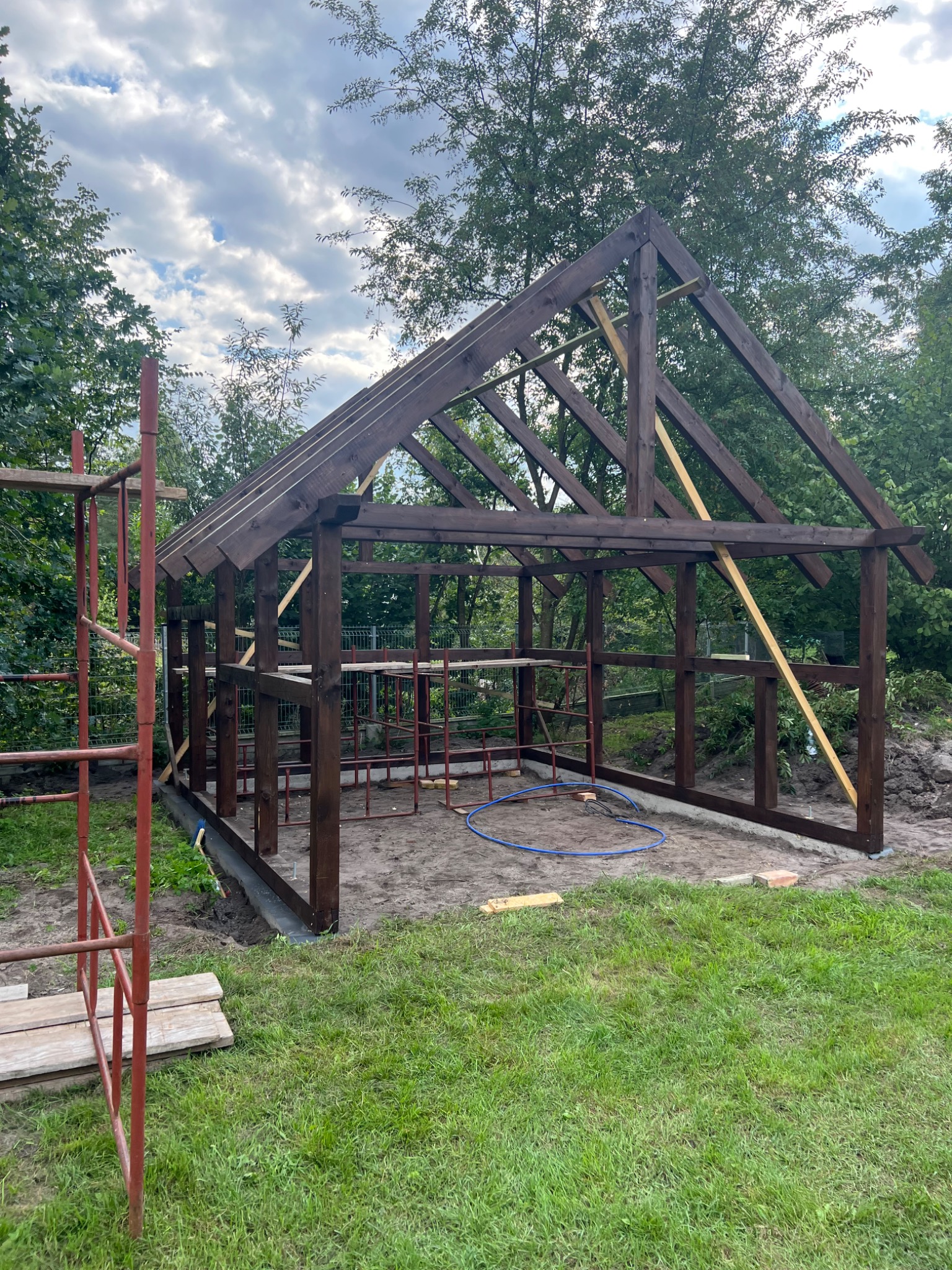 Konstrukcja drewniana altany w trakcie budowy, widoczne rusztowanie i elementy więźby dachowej na tle zieleni i pochmurnego nieba.