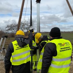 Metis Intech sp. z o.o. - Montaż konstrukcji wsporczej pod panele. Ekipa w żółtych kaskach i odblaskowych kamizelkach pracuje na zewnątrz przy pochmurnej pogodzie. Widoczne elementy maszyn.