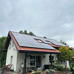 Metis Intech sp. z o.o. - Biały dom z czerwoną dachówką i panelami słonecznymi. Na werandzie kwiaty w donicach, siedząca osoba. Pochmurne niebo, drzewa w tle.