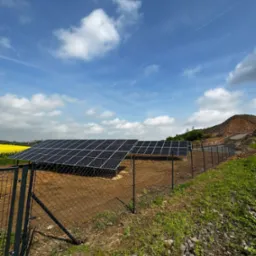 Instalacja paneli słonecznych za ogrodzeniem z siatki, na tle błękitnego nieba z chmurami i pagórkowatego terenu z żółtymi polami w oddali.