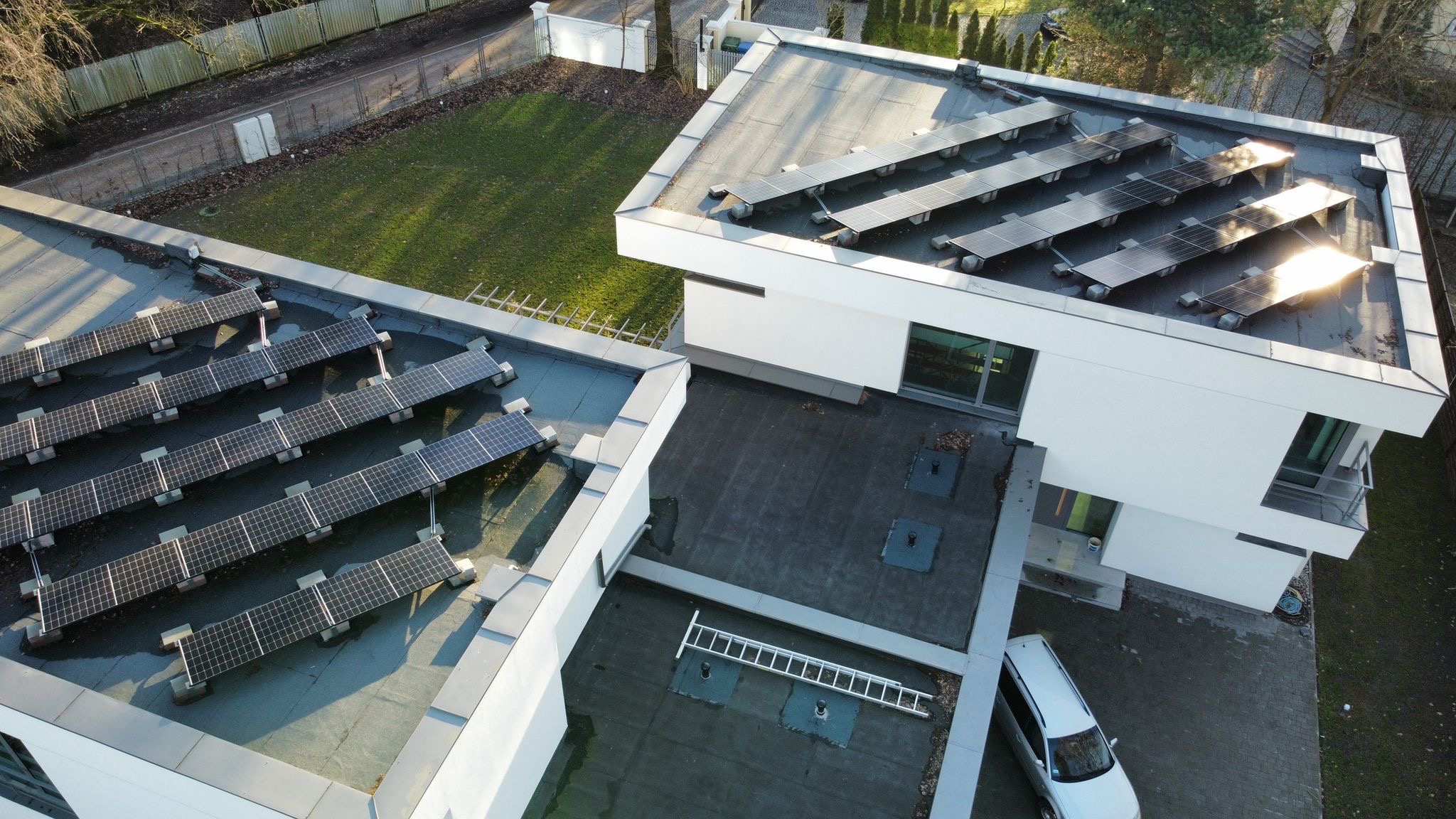 Widok z góry na nowoczesny dom z panelami słonecznymi na płaskim dachu. Drabina leży obok, a obok domu stoi srebrny samochód. Zieleń trawnika w tle.