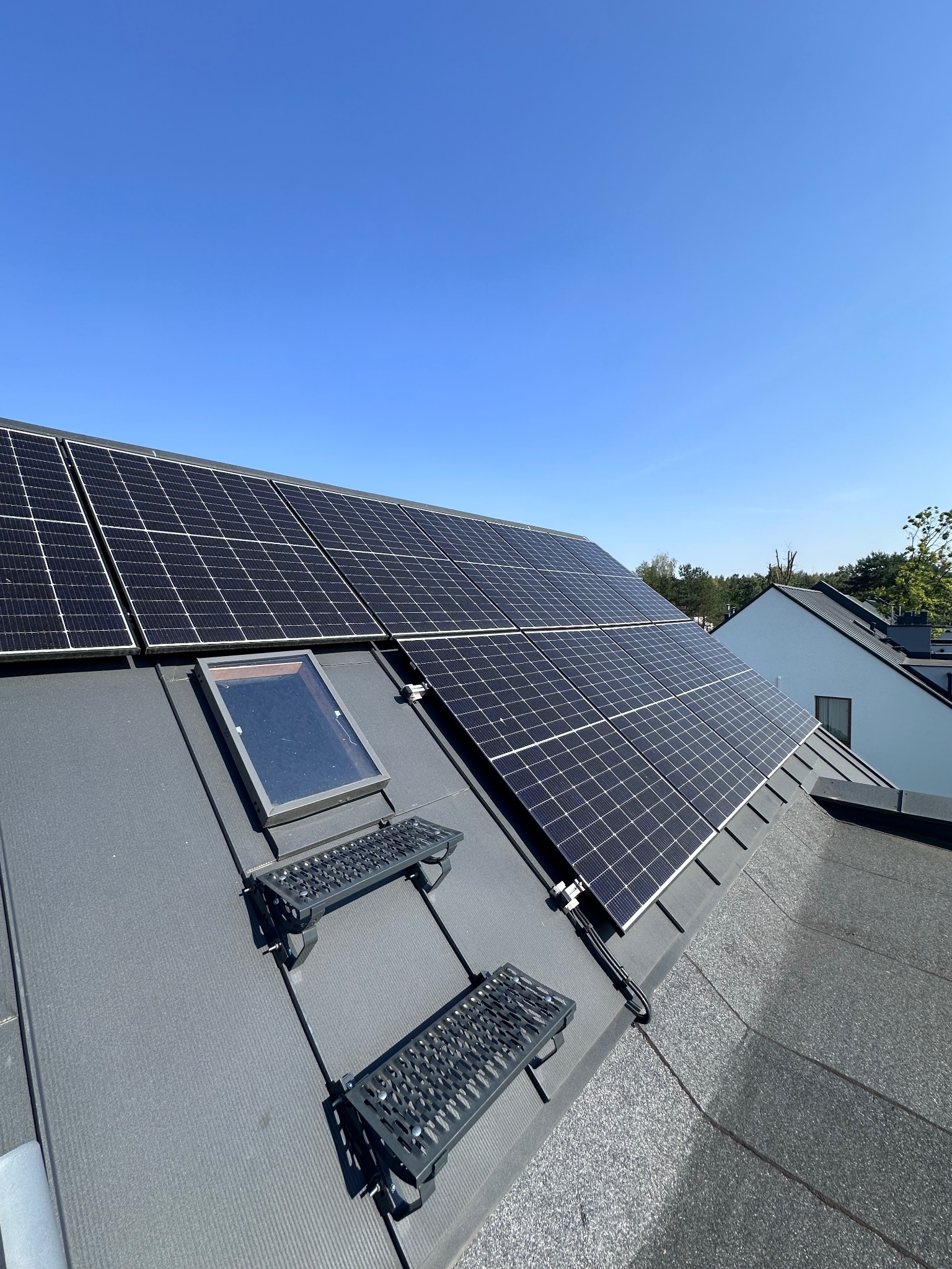 Panele fotowoltaiczne na dachu z szarym pokryciem, widoczne okno dachowe i metalowe stopnie. Błękitne niebo w tle.