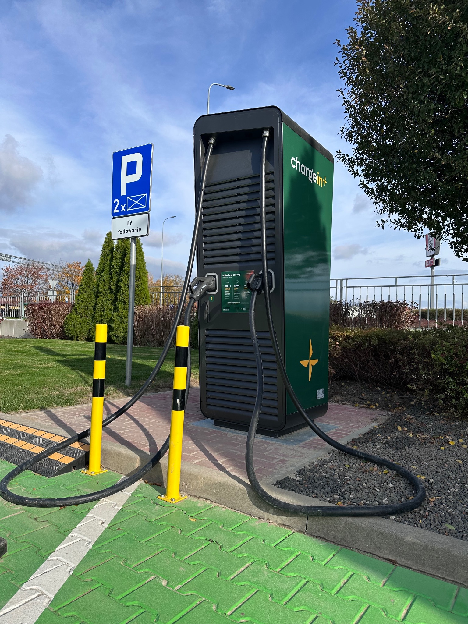 Stacja ładowania pojazdów elektrycznych Chargein+ na zielono-szarym parkingu, z żółto-czarnymi słupkami zabezpieczającymi i znakiem parkingowym w tle.