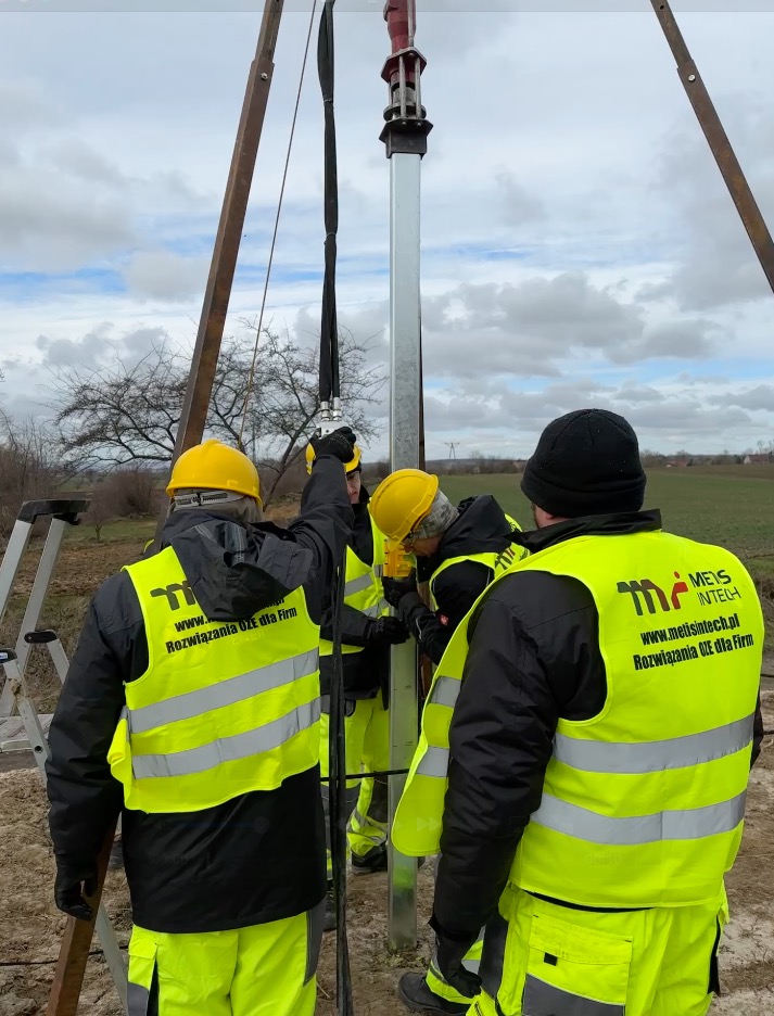 Montaż konstrukcji wsporczej pod panele. Ekipa w żółtych kaskach i odblaskowych kamizelkach pracuje na zewnątrz przy pochmurnej pogodzie. Widoczne elementy maszyn.