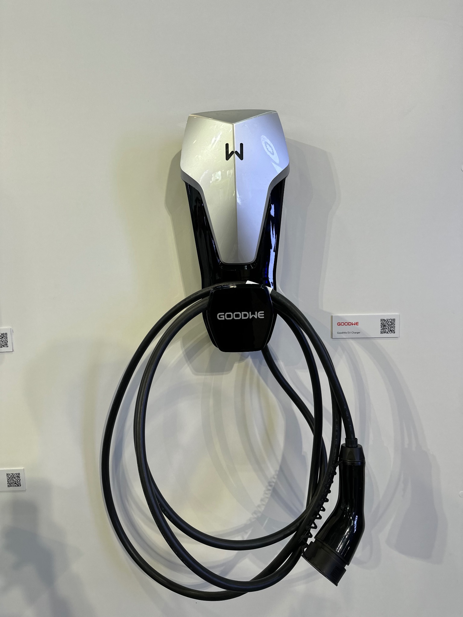Stacja ładowania EV GoodWe na ścianie, z kablem zwiniętym w pętle. Widoczne oznaczenia producenta i kod QR. Nowoczesny design, stonowana kolorystyka.