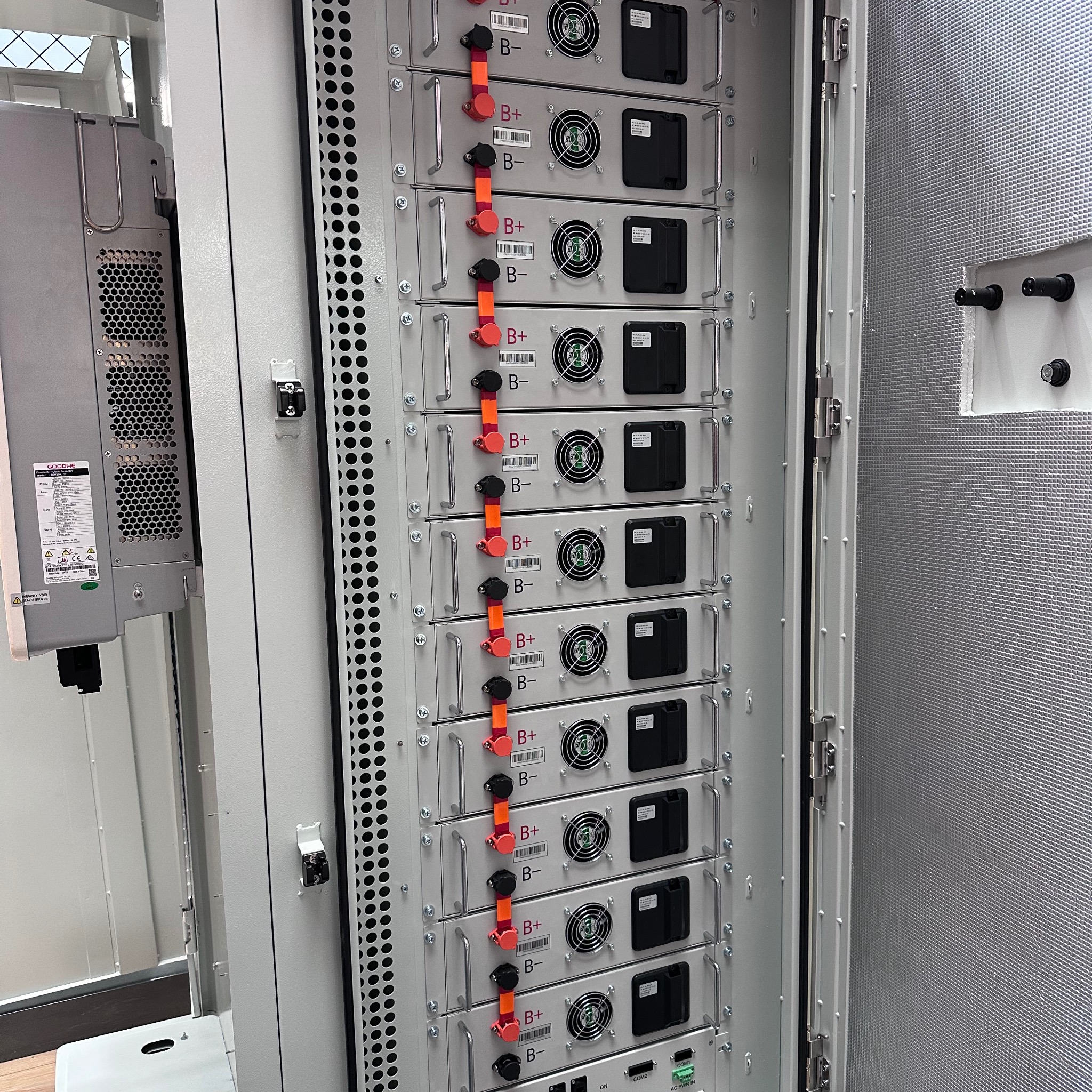 Wnętrze szafy z modułowymi magazynami energii, widoczne liczne baterie z oznaczeniami biegunów B+ i B-, wentylatory chłodzące i elementy instalacji elektrycznej.