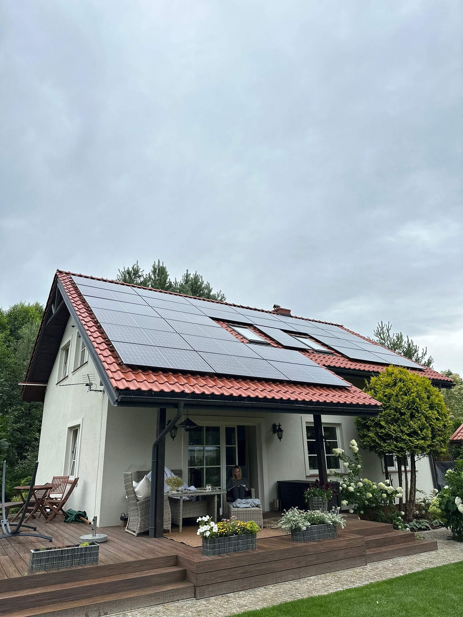 Biały dom z czerwoną dachówką i panelami słonecznymi. Na werandzie kwiaty w donicach, siedząca osoba. Pochmurne niebo, drzewa w tle.
