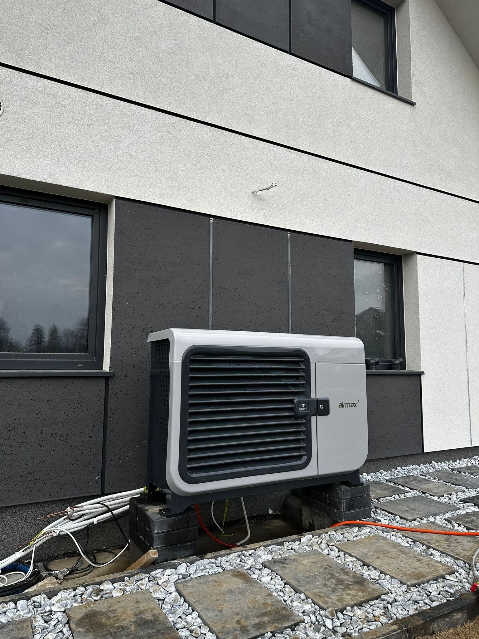 Szara pompa ciepła Airmax na zewnątrz nowoczesnego domu z elewacją w odcieniach szarości i białego, na tle kamiennej ścieżki i przewodów.