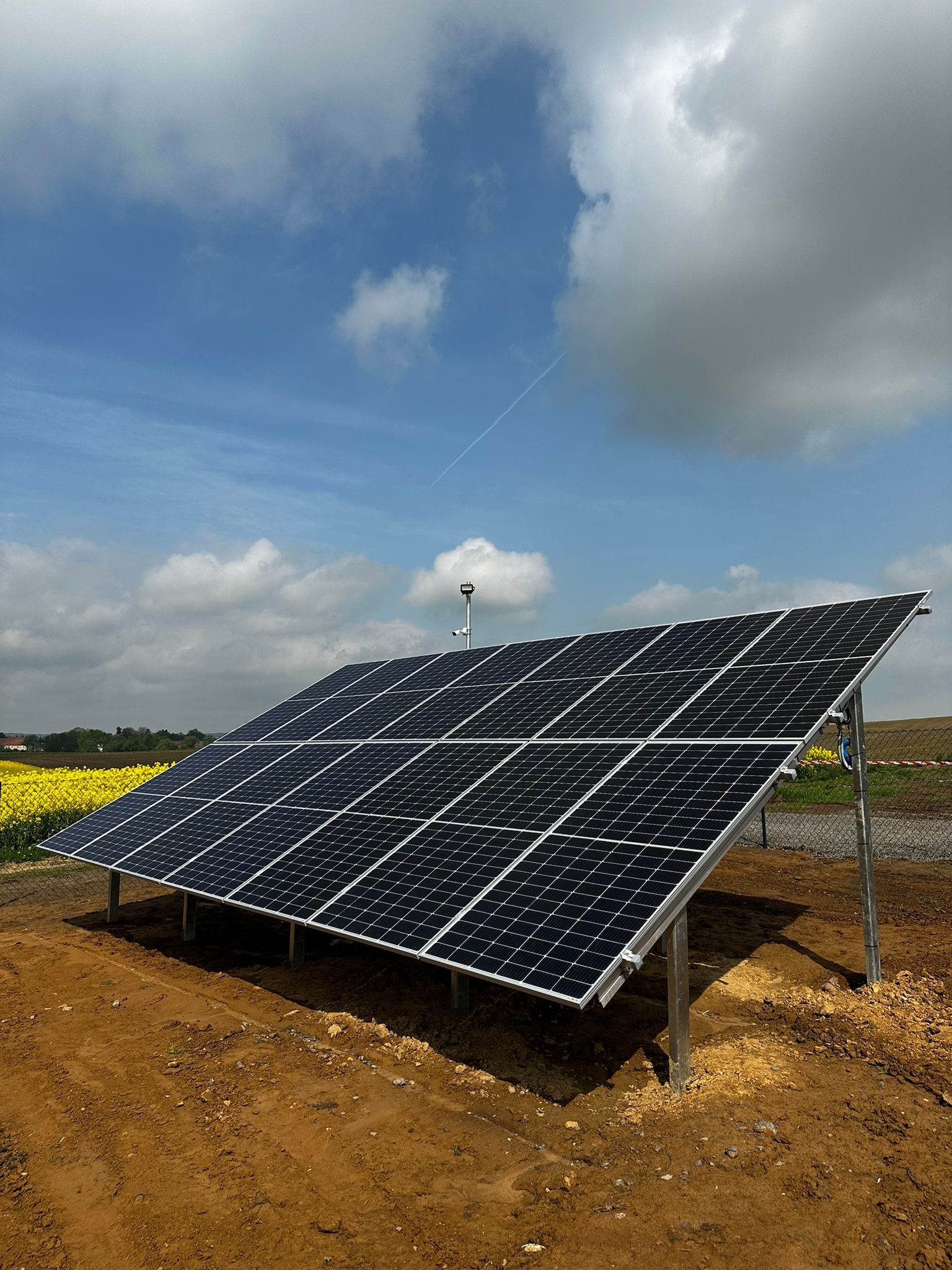 Panel fotowoltaiczny na metalowej konstrukcji, ustawiony na ziemi. W tle pole rzepaku i błękitne niebo z chmurami. Widoczny biały ślad po samolocie.