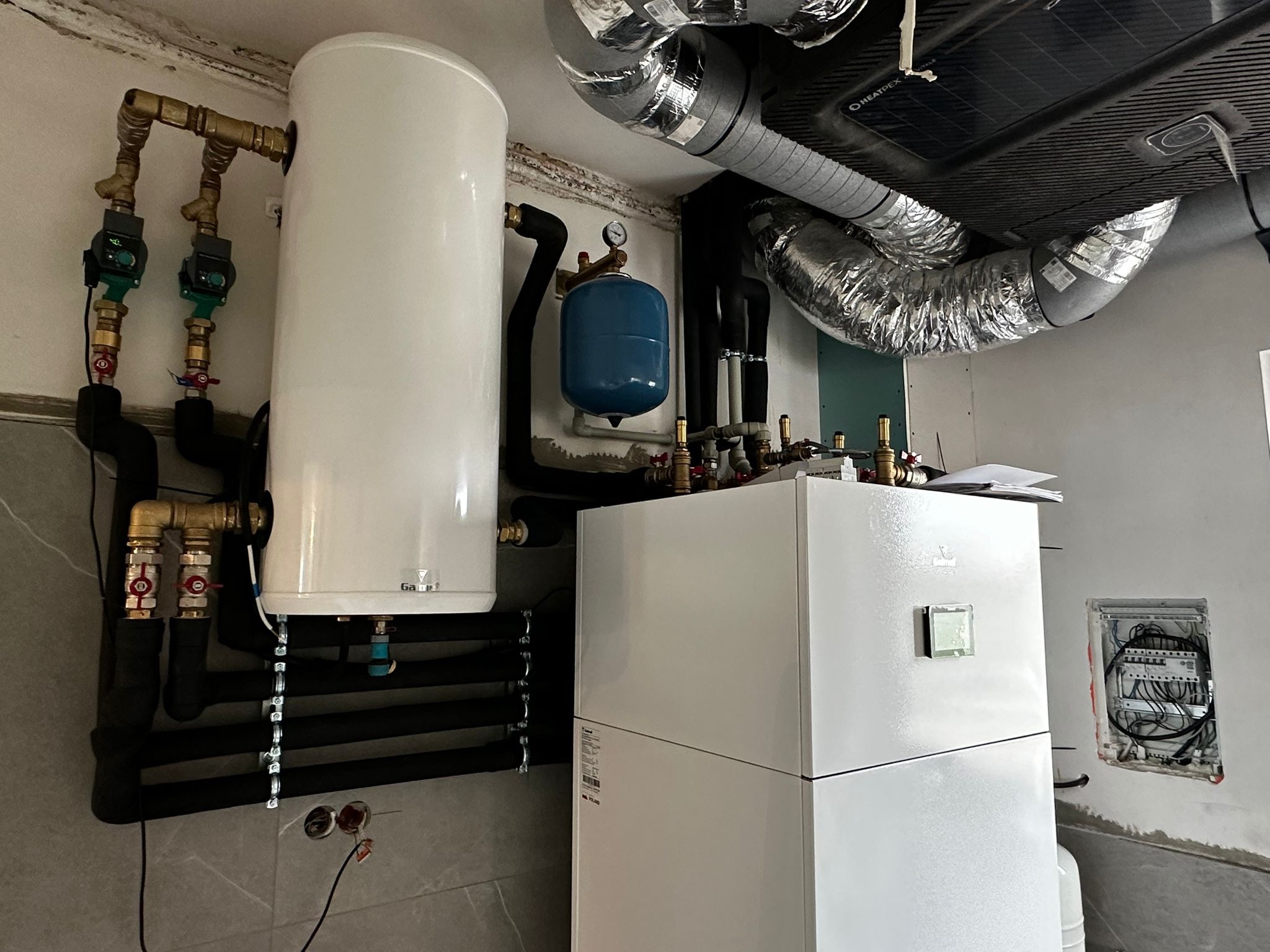 Biały zbiornik ciepłej wody użytkowej i pompa ciepła w kotłowni, z widoczną instalacją hydrauliczną i elektryczną, fragment wentylacji mechanicznej.