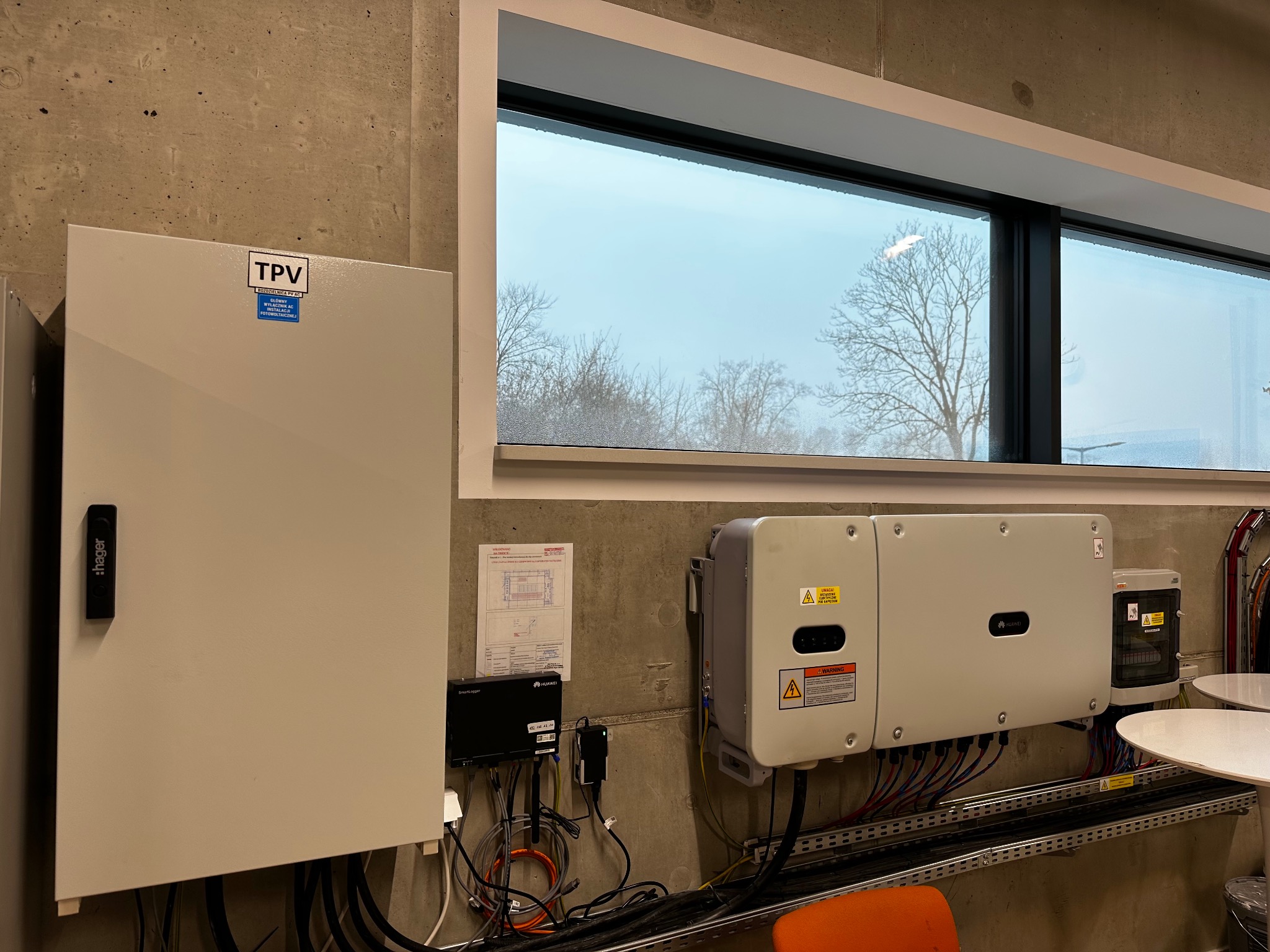 Instalacja fotowoltaiczna wewnątrz budynku: rozdzielnica TPV, inwertery Huawei z okablowaniem na tle betonowej ściany i okna. Widoczne elementy instalacji elektrycznej.