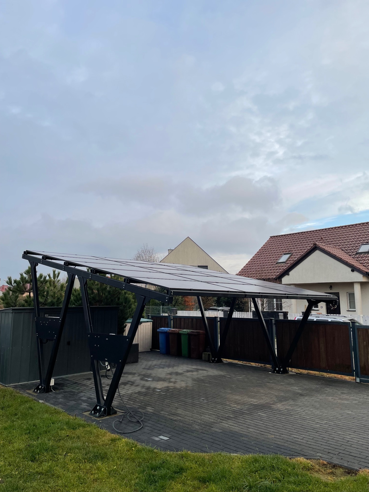 Nowoczesna wiata fotowoltaiczna na posesji, z czarną konstrukcją i panelami słonecznymi. Widoczne domy w tle i kostka brukowa na podjeździe.