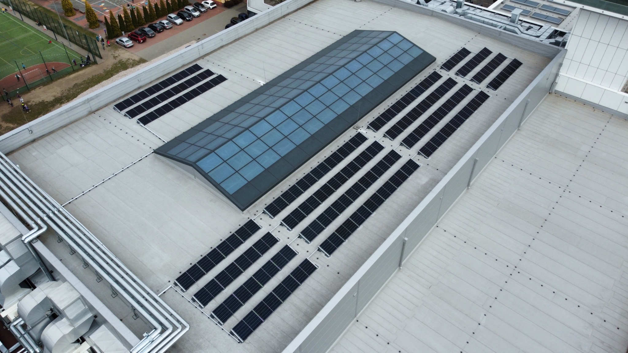 Widok z góry na płaski dach budynku z zainstalowanymi panelami fotowoltaicznymi w rzędach, obok świetlika dachowego. W tle boisko i parking.