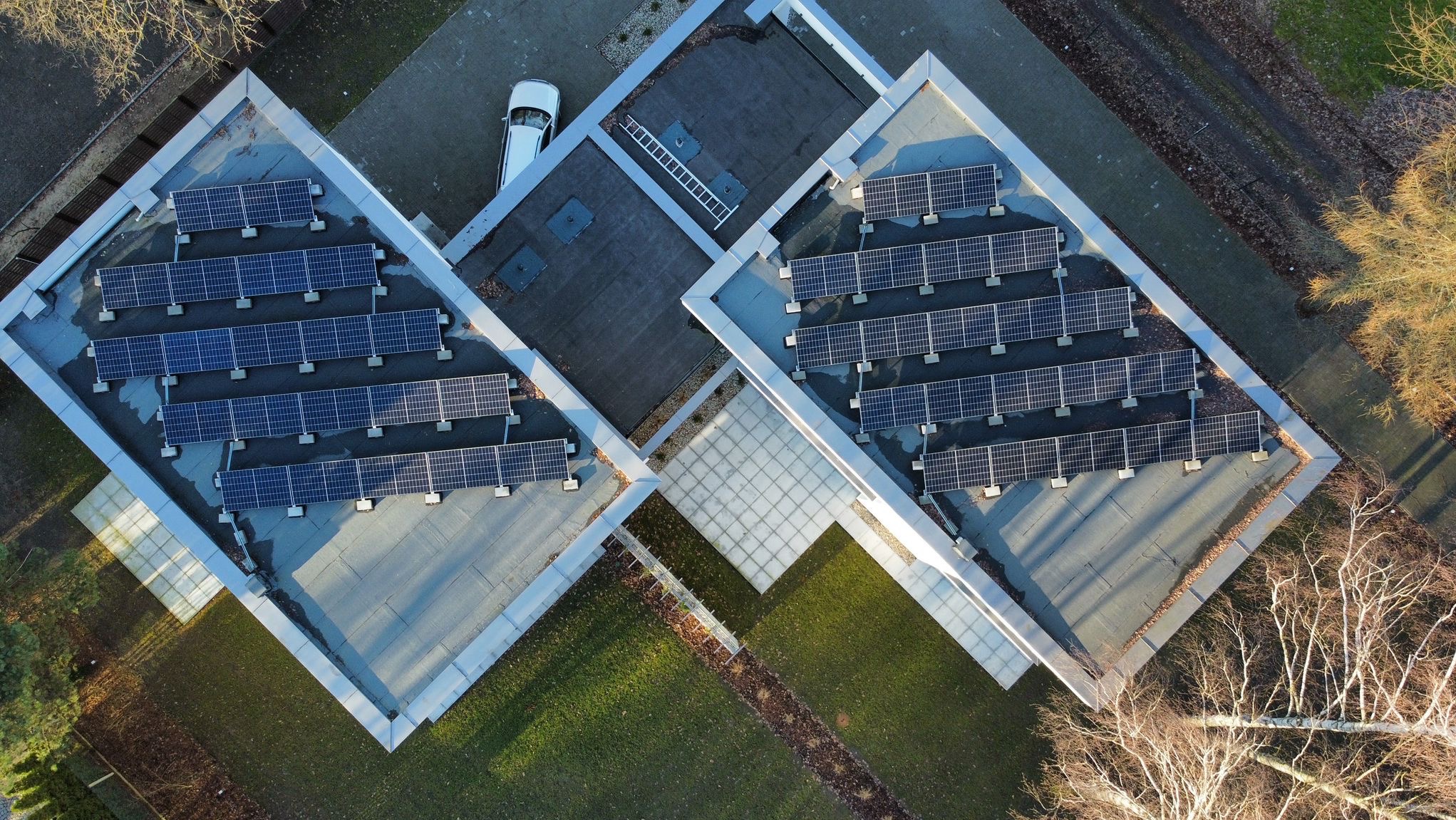 Widok z góry na dach budynku z panelami słonecznymi, obok trawnik i drabina. Ujęcie pokazuje instalację fotowoltaiczną w otoczeniu zieleni.