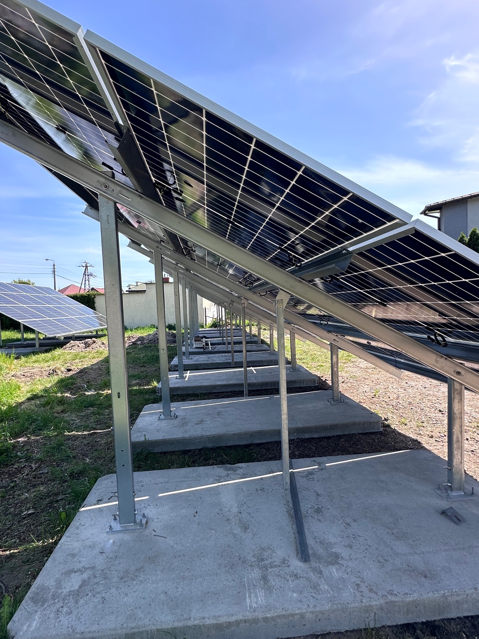 Rząd paneli słonecznych zamontowanych na betonowych podstawach, widok z dołu, z kolejnym rzędem paneli w tle i fragmentem budynku. Błękitne niebo.