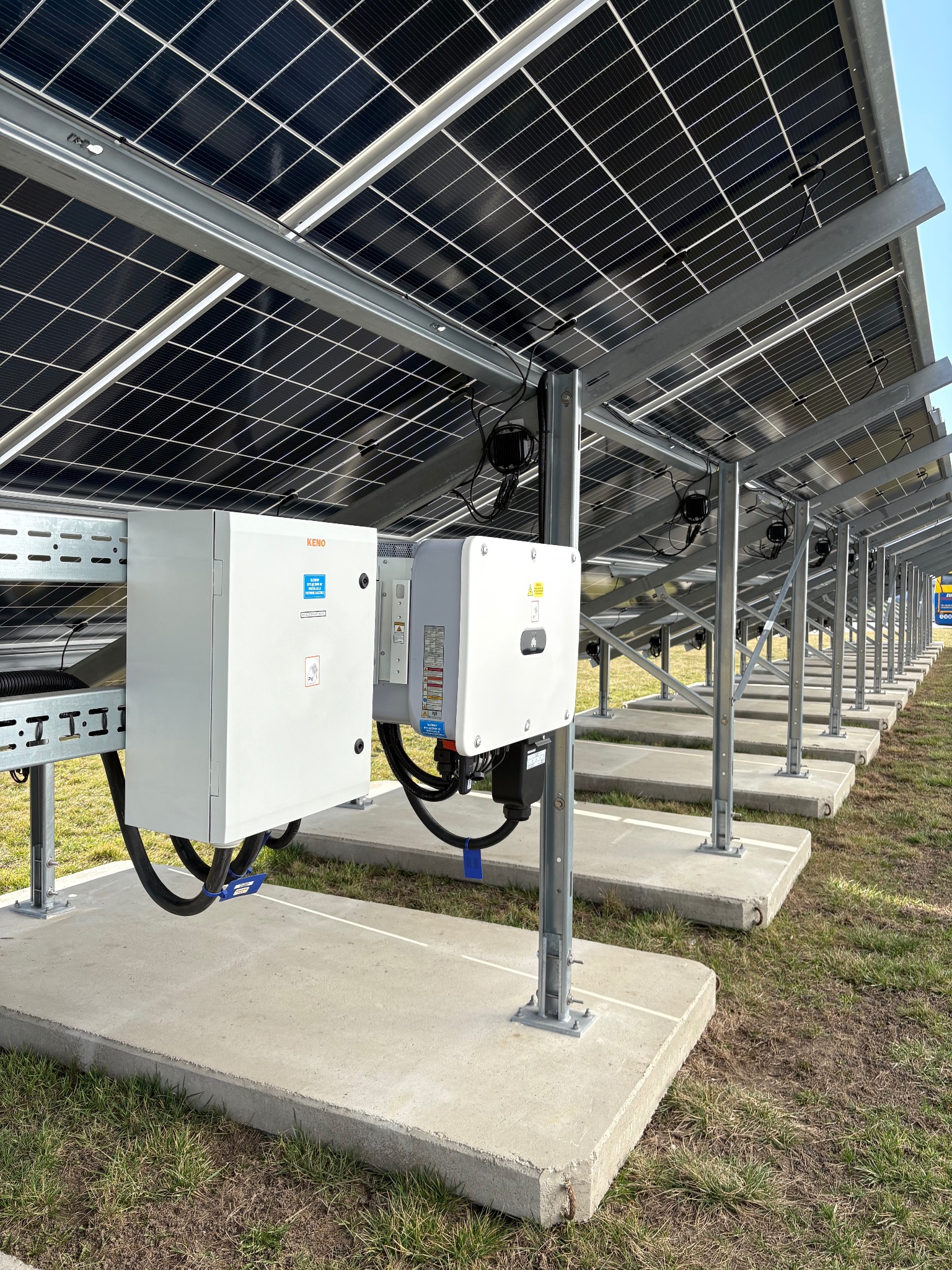 Rząd paneli słonecznych z widocznymi inwerterami i konstrukcją wsporczą, zamontowanych na betonowych podstawach na trawiastym terenie. Perspektywa od dołu.