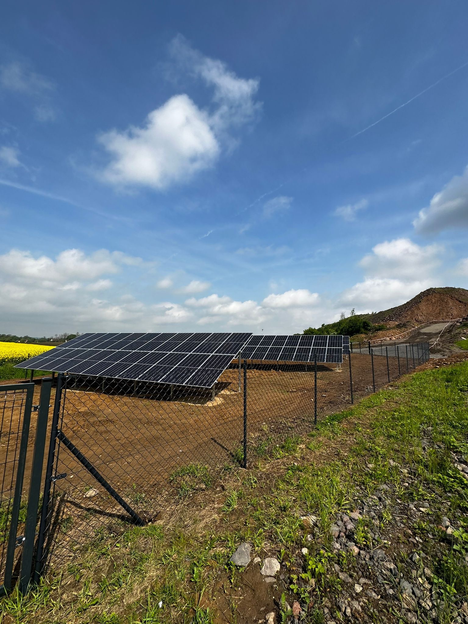 Instalacja paneli słonecznych za ogrodzeniem z siatki, na tle błękitnego nieba z chmurami i pagórkowatego terenu z żółtymi polami w oddali.