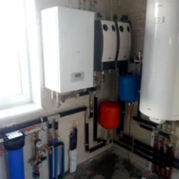 Kompletna instalacja grzewcza w kotłowni: biały piec gazowy, dwa zasobniki ciepłej wody z czarnymi obudowami, filtry wody z niebieskimi obudowami, zbiorniki wyrównawcze w kolorze niebieskim...