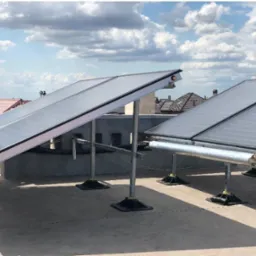 Kolektory słoneczne zamontowane na płaskim dachu budynku mieszkalnego, widoczne rury i mocowania, w tle antena satelitarna i zabudowa miejska pod błękitnym niebem z chmurami.