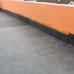 Wykończony płaski dach z papy termozgrzewalnej, pomarańczowa attyka z widocznymi śladami po butach, w tle zabudowania mieszkalne.