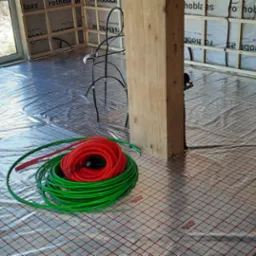 Rozwinięta folia grzewcza na podłodze w trakcie montażu ogrzewania podłogowego, widoczna rolka zielonych rur na wózku oraz zwój rur czerwonych i zielonych, ściany w surowym stanie.