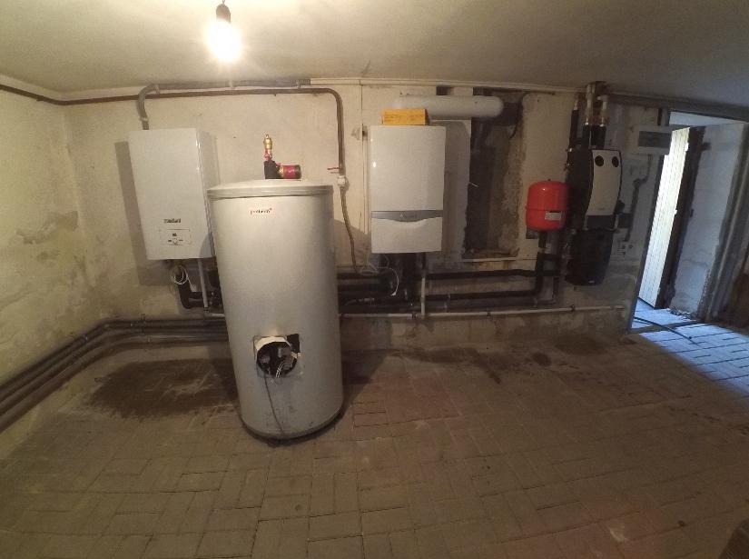 Piwnica z dwoma piecami gazowymi Vaillant, zasobnikiem wody i instalacją hydrauliczną, widoczne rury grzewcze i elementy montażowe.