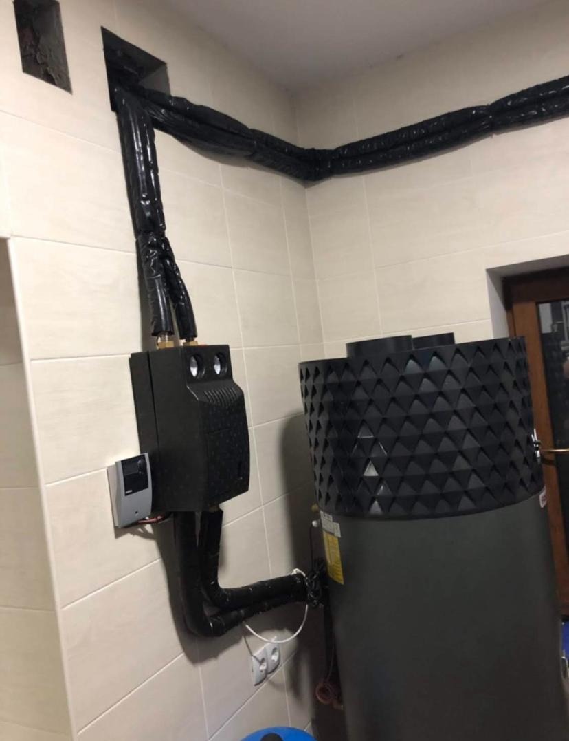 Czarny moduł sterujący i zbiornik ciepłej wody połączone izolowanymi przewodami w pomieszczeniu z jasnymi płytkami; widoczne gniazdka elektryczne.