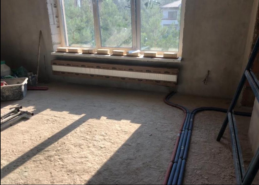 Surowy stan wnętrza podczas montażu instalacji ogrzewania podłogowego, widoczne rury grzewcze rozchodzące się na posadzce, grzejnik w kartonie pod oknem, drabina i narzędzia.