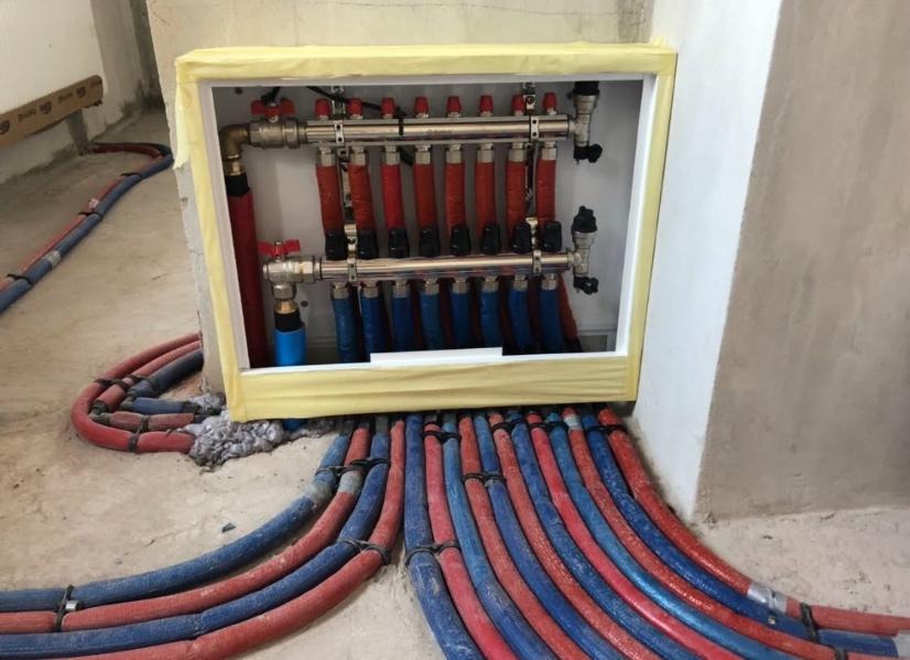 Rozdzielacz ogrzewania podłogowego w obudowie, z widocznymi rurami w kolorach czerwonym i niebieskim, prowadzącymi w podłogę, zabezpieczony taśmą malarską.