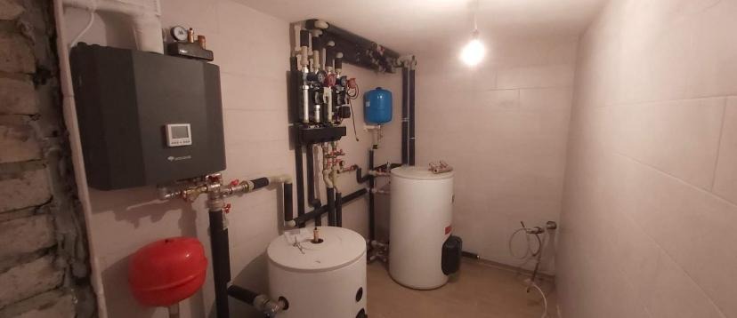 Węzeł cieplny z dwoma białymi zasobnikami ciepłej wody, szarą centralą sterującą i licznymi rurami w kotłowni w Niepołomicach.