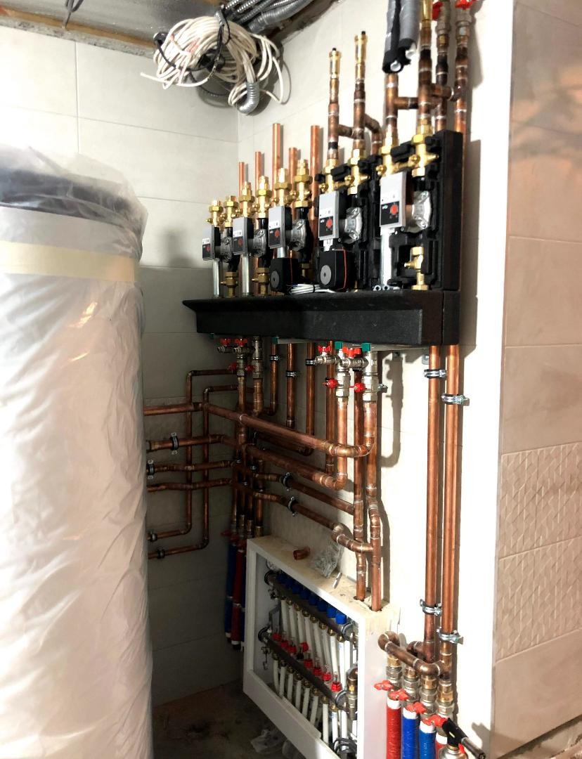 Rozdzielacz ogrzewania podłogowego z rurami miedzianymi i osprzętem, widoczny zasobnik ciepłej wody w folii ochronnej, instalacja w pomieszczeniu z białymi płytkami.