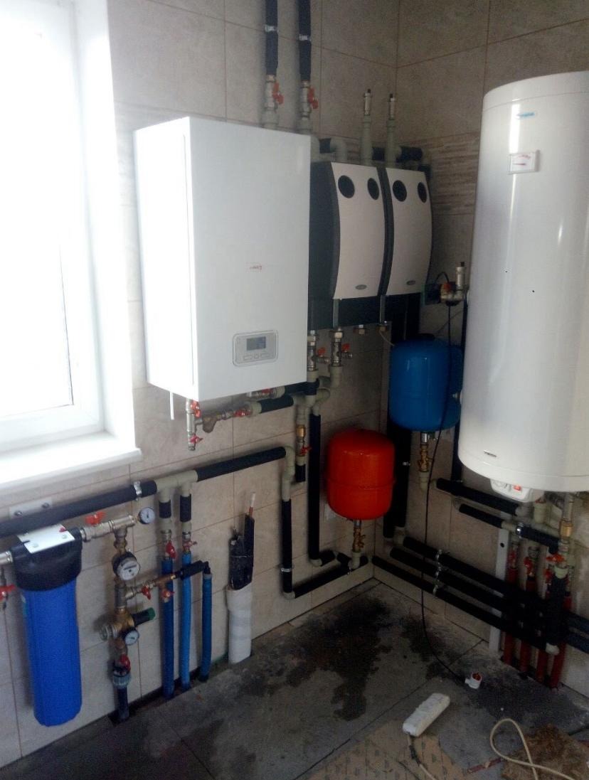 Kompletna instalacja grzewcza w kotłowni: biały piec gazowy, dwa zasobniki ciepłej wody z czarnymi obudowami, filtry wody z niebieskimi obudowami, zbiorniki wyrównawcze w kolorze niebieskim...