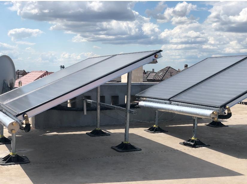 Kolektory słoneczne zamontowane na płaskim dachu budynku mieszkalnego, widoczne rury i mocowania, w tle antena satelitarna i zabudowa miejska pod błękitnym niebem z chmurami.