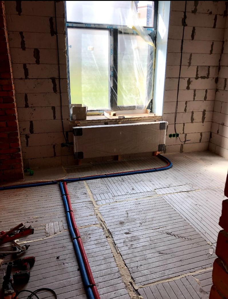 Montaż ogrzewania podłogowego w surowym stanie: widoczne rury grzewcze w kolorze niebieskim i czerwonym ułożone na przygotowanym podłożu, ściany z betonu komórkowego i fragment ściany z czerwonej...