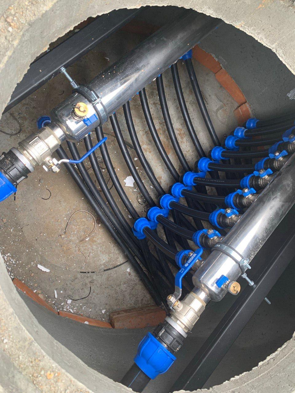 Rozdzielacz z czarnymi rurami i niebieskimi złączkami w betonowym kręgu studni, widoczne zawory kulowe i fragmenty cegieł.