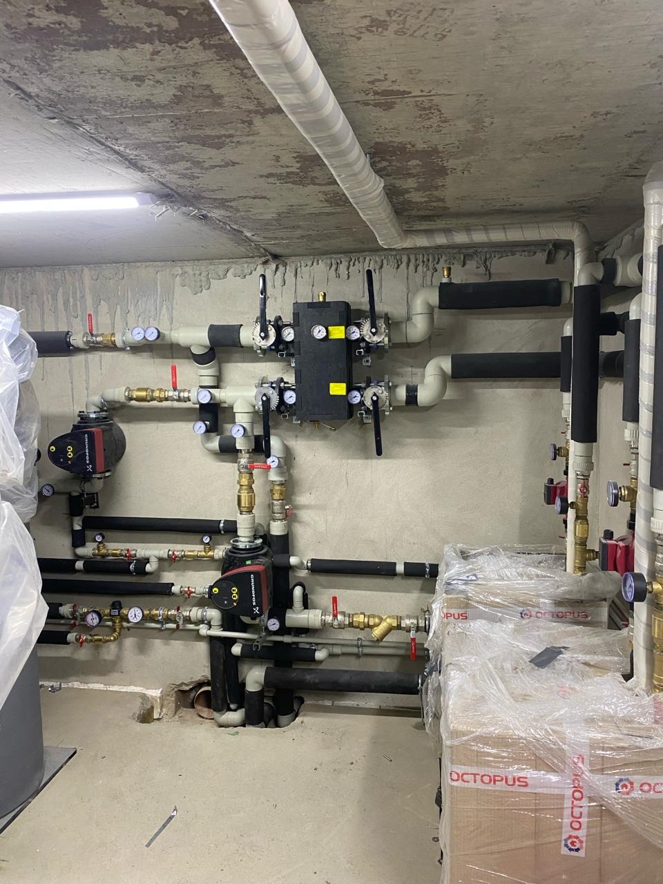 Złożona instalacja rur, zaworów, manometrów i pomp obiegowych, z czarną skrzynką rozdzielczą i owiniętymi rurami, zamontowana na szarej ścianie w kotłowni, z kartonami w folii na podłodze.
