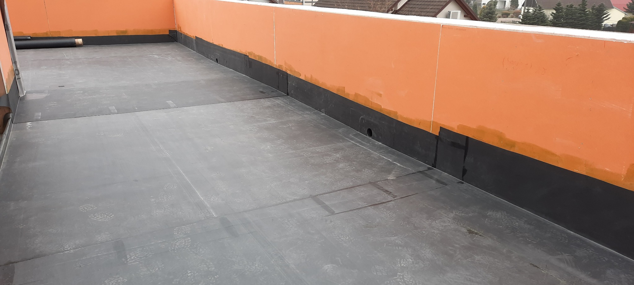 Wykończony płaski dach z papy termozgrzewalnej, pomarańczowa attyka z widocznymi śladami po butach, w tle zabudowania mieszkalne.