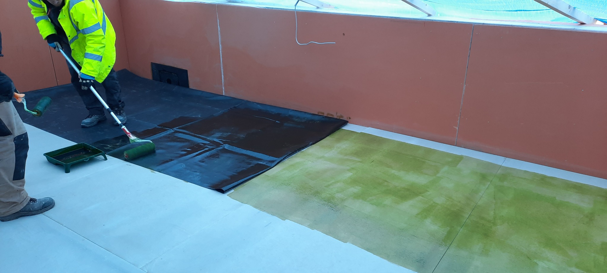Hydroizolacja podłogi wewnątrz budynku: robotnik w odblaskowej kurtce nanosi wałkiem czarną powłokę na folię, obok widoczna zielona warstwa hydroizolacyjna i kuweta z farbą.