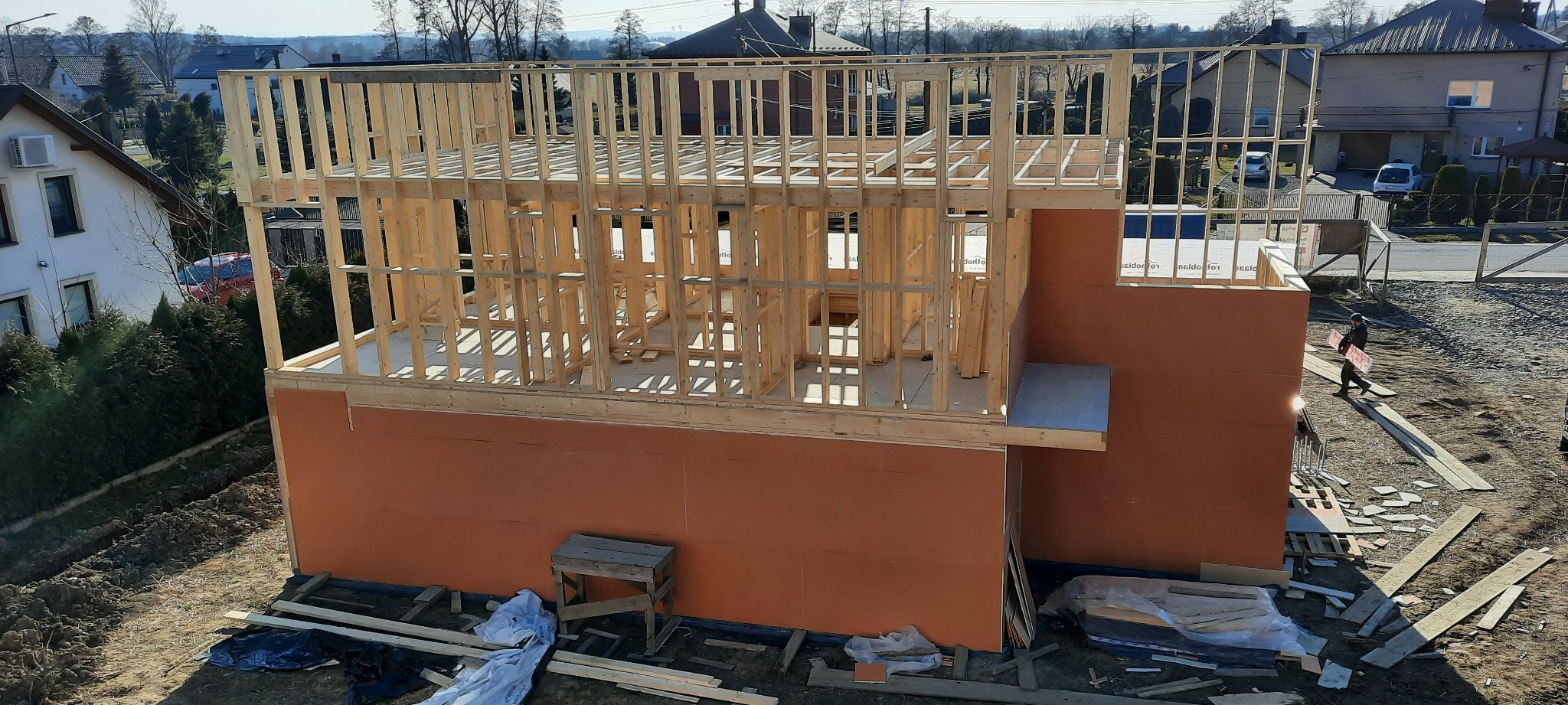 Budowa domu szkieletowego: widoczna konstrukcja drewniana z pomarańczową płytą OSB, pracownik przenosi materiały budowlane na zewnątrz.