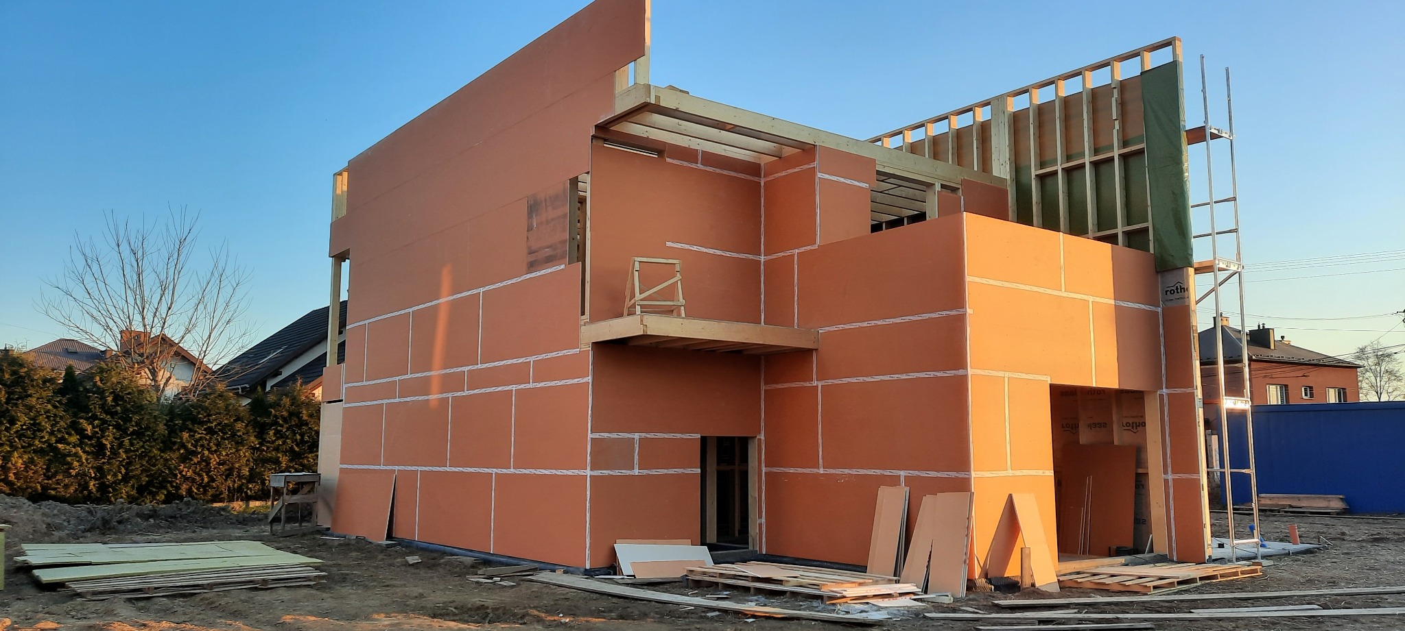 Surowy stan domu szkieletowego z pomarańczowymi panelami izolacyjnymi i drewnianą konstrukcją, widoczne rusztowania i materiały budowlane na placu budowy.