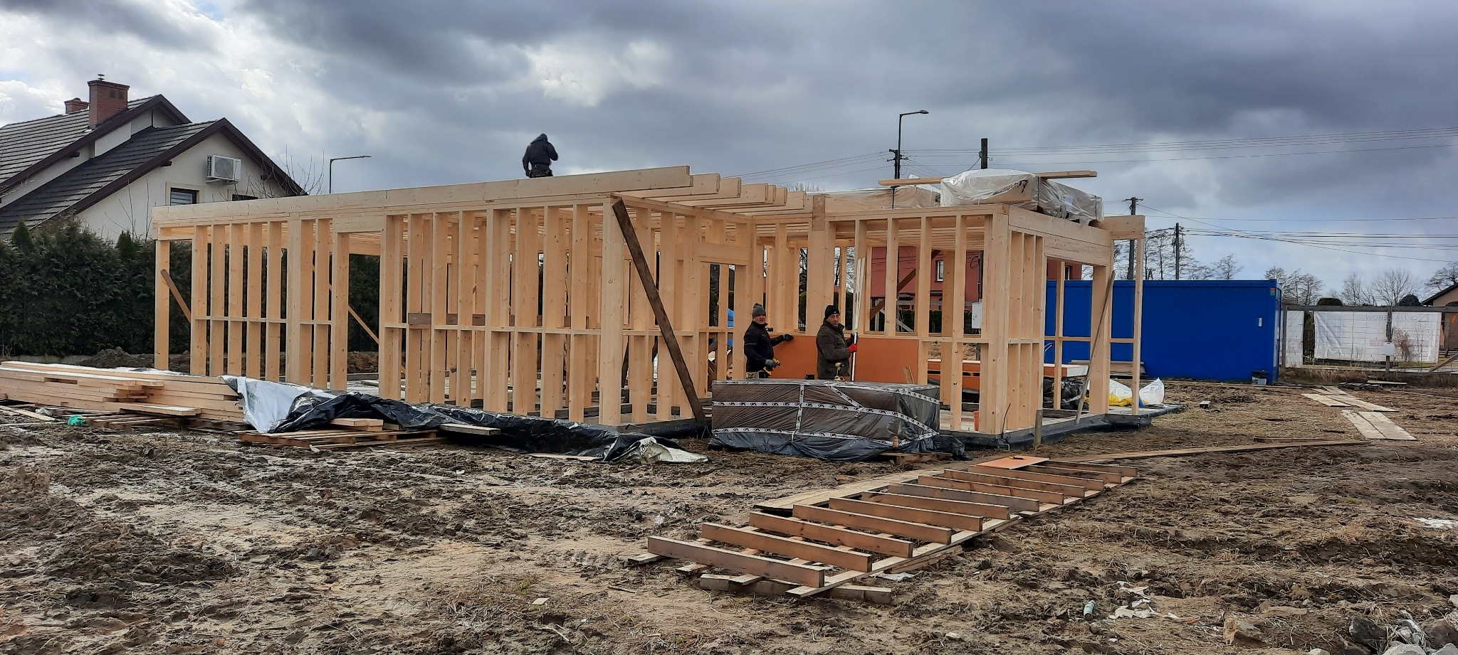 Wznoszenie szkieletu drewnianego domu: trzech pracowników montuje ściany z drewnianych belek na błotnistym placu budowy, w tle dom jednorodzinny i błękitny kontener.