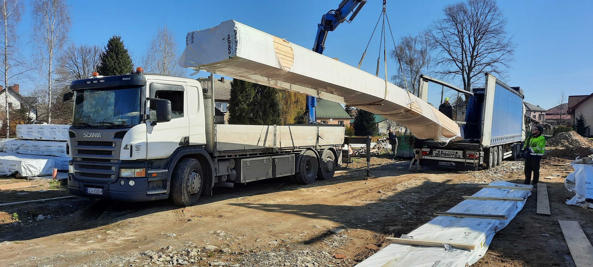 Rozładunek dźwigiem modułów domu szkieletowego z ciężarówki na placu budowy, widoczni pracownicy w odblaskowych kamizelkach.