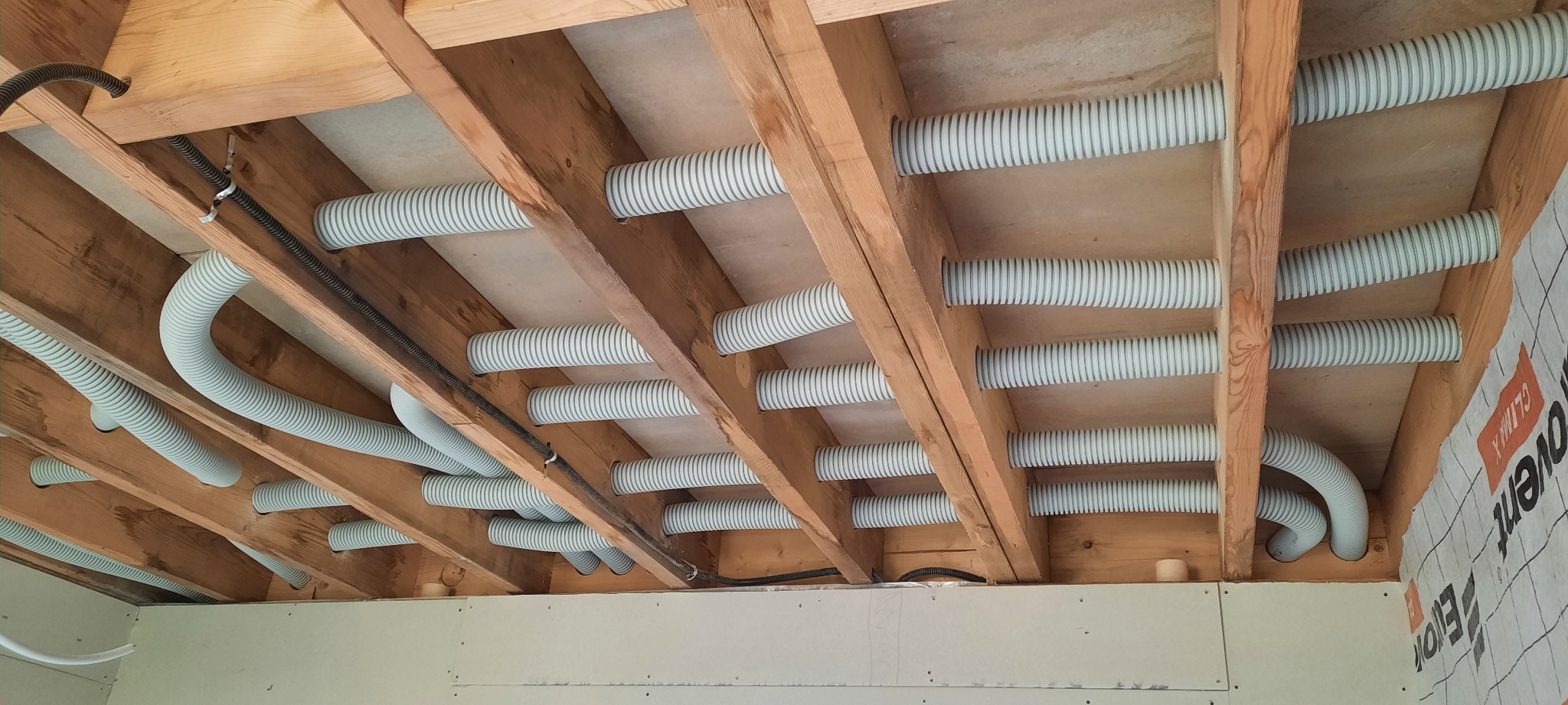 Szare, elastyczne rury wentylacyjne ułożone między drewnianymi belkami stropu podczas montażu systemu wentylacji mechanicznej z rekuperacją w nowym domu.