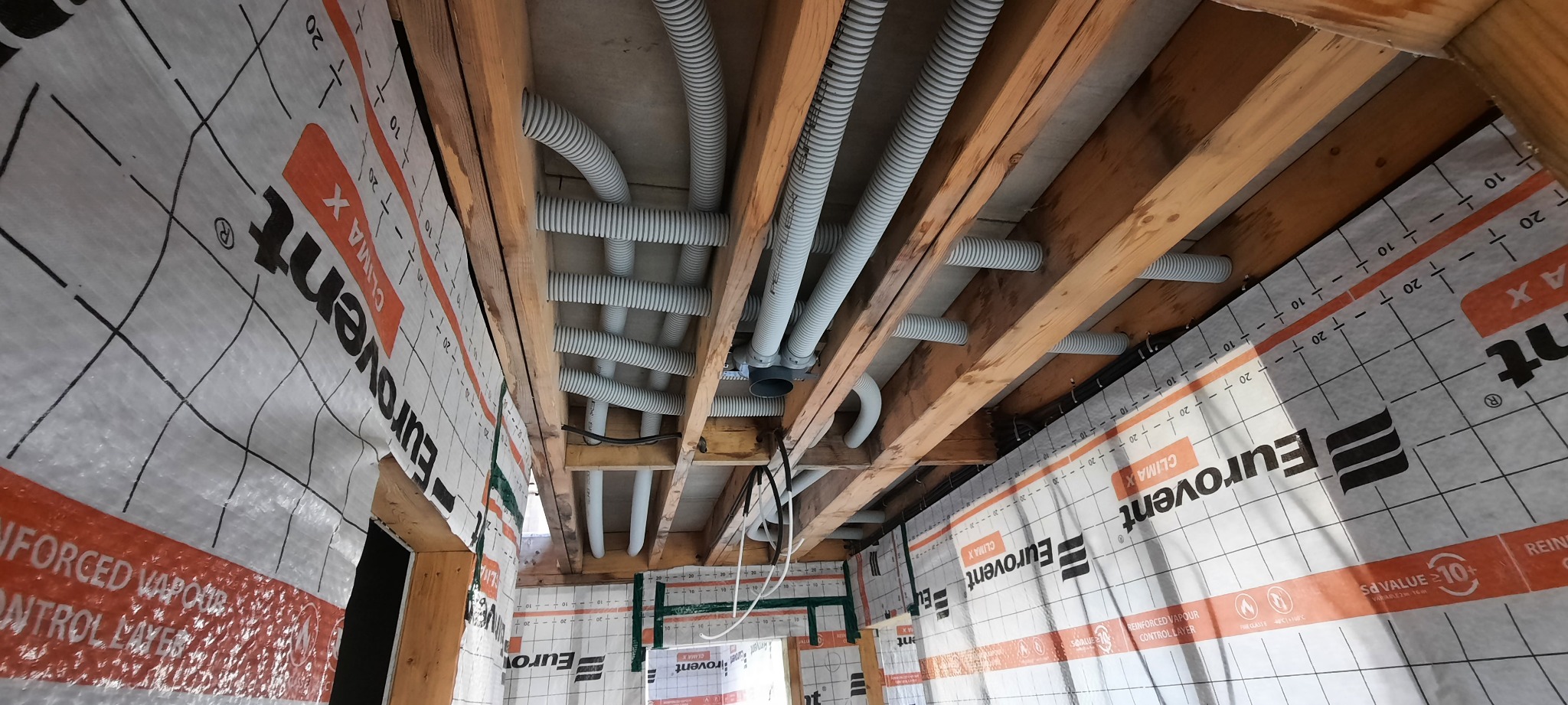 Montaż instalacji wentylacyjnej z rurami karbowanymi pod stropem, widoczne drewniane belki konstrukcyjne i folia paroizolacyjna Eurovent Clima X na ścianach.