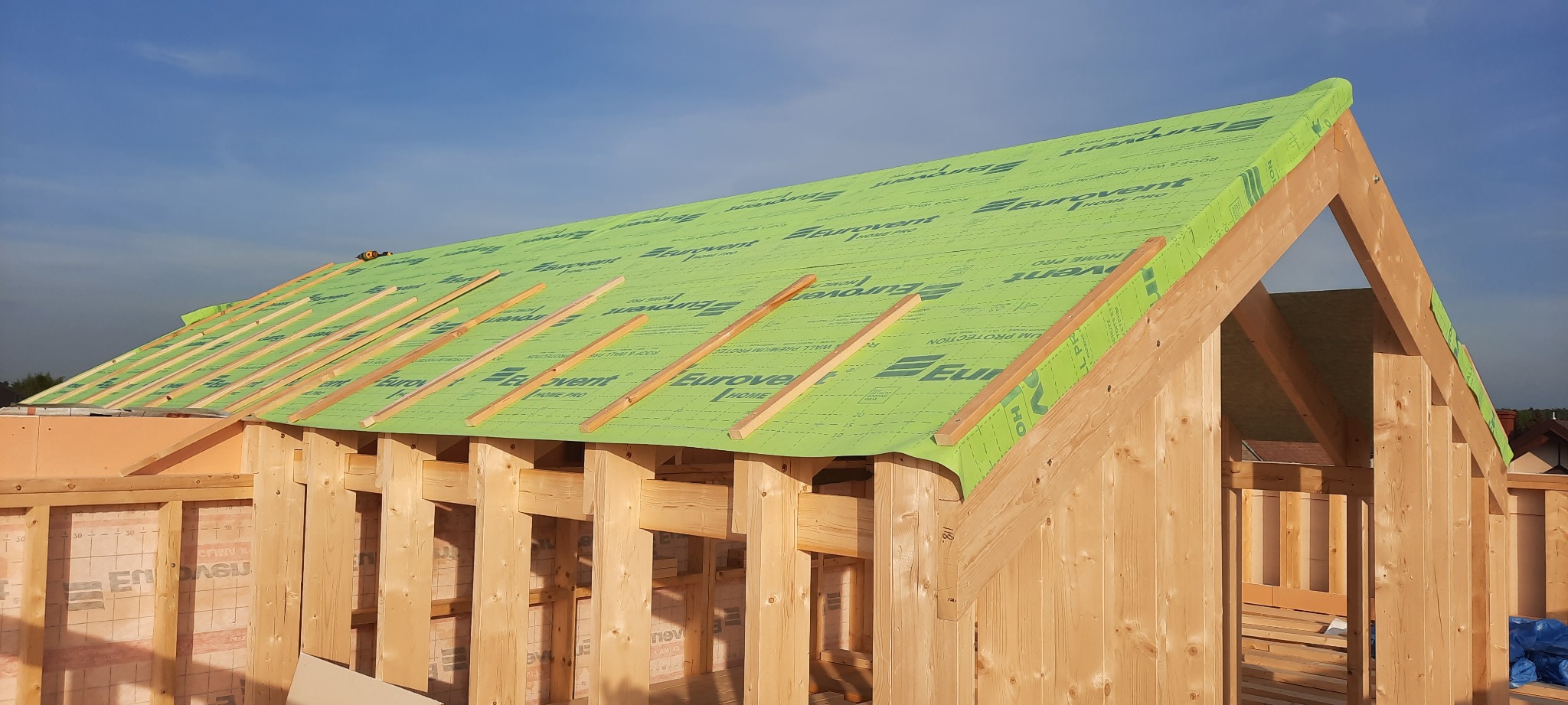 Konstrukcja drewniana domu w trakcie budowy z widoczną membraną dachową Eurovent i elementami więźby dachowej, jasne drewno, niebieskie niebo w tle.
