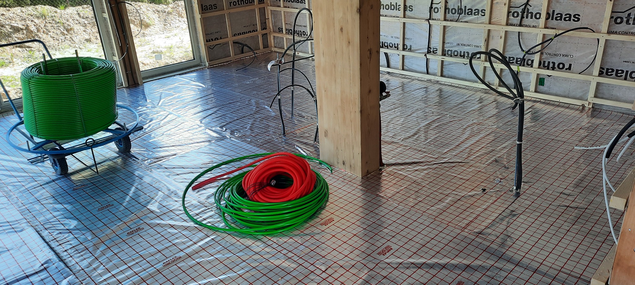 Rozwinięta folia grzewcza na podłodze w trakcie montażu ogrzewania podłogowego, widoczna rolka zielonych rur na wózku oraz zwój rur czerwonych i zielonych, ściany w surowym stanie.