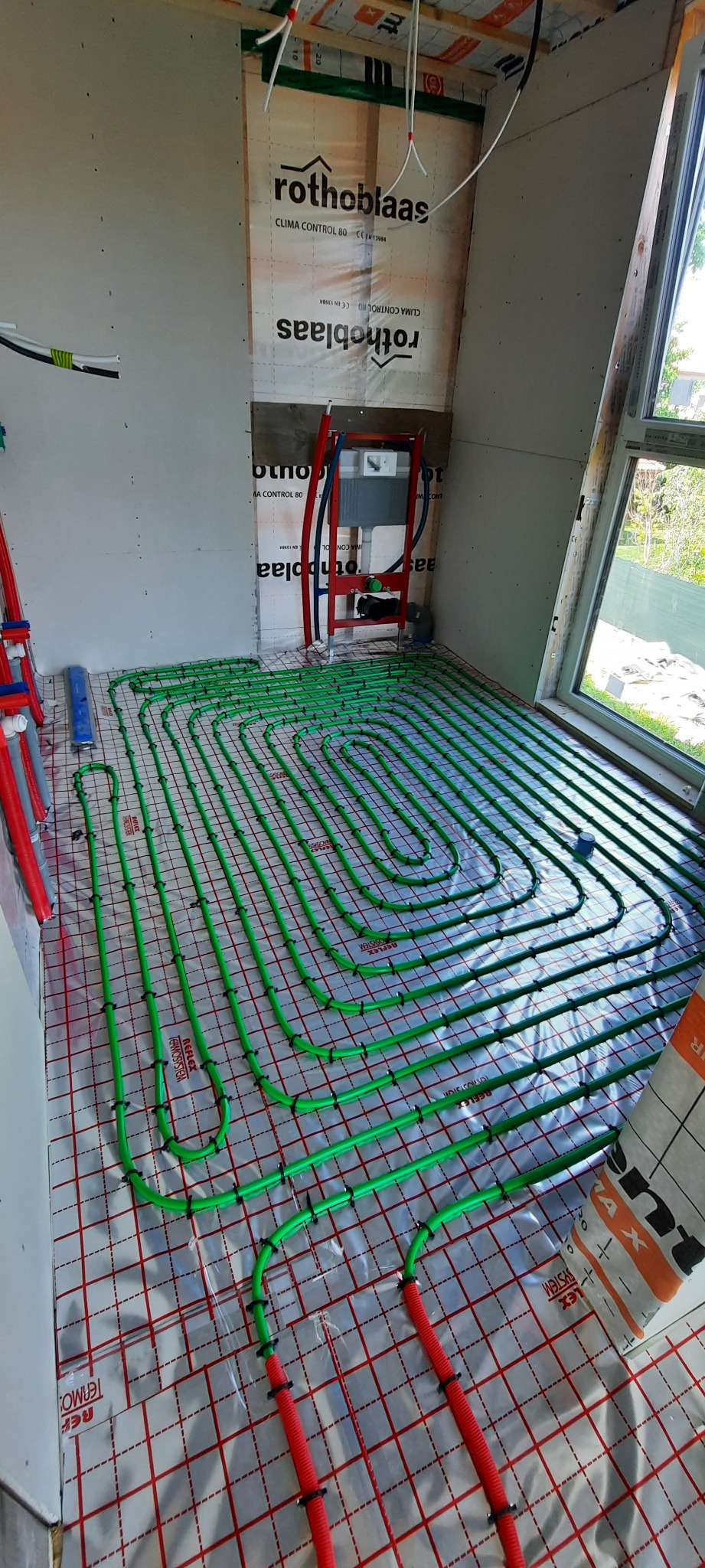 Montaż zielonych rur ogrzewania podłogowego na izolacji z czerwoną siatką w surowym wnętrzu, widoczne elementy instalacji sanitarnej i okno.