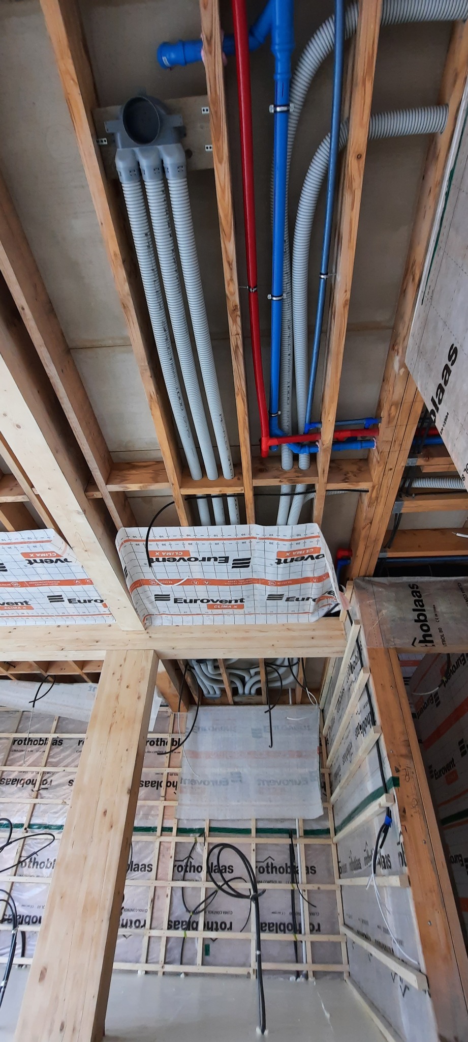 Wnętrze domu w trakcie budowy z widocznymi instalacjami wentylacyjnymi (szare, elastyczne rury) oraz instalacjami wodnymi (czerwone i niebieskie rury) biegnącymi pod drewnianym stropem, częściowo...