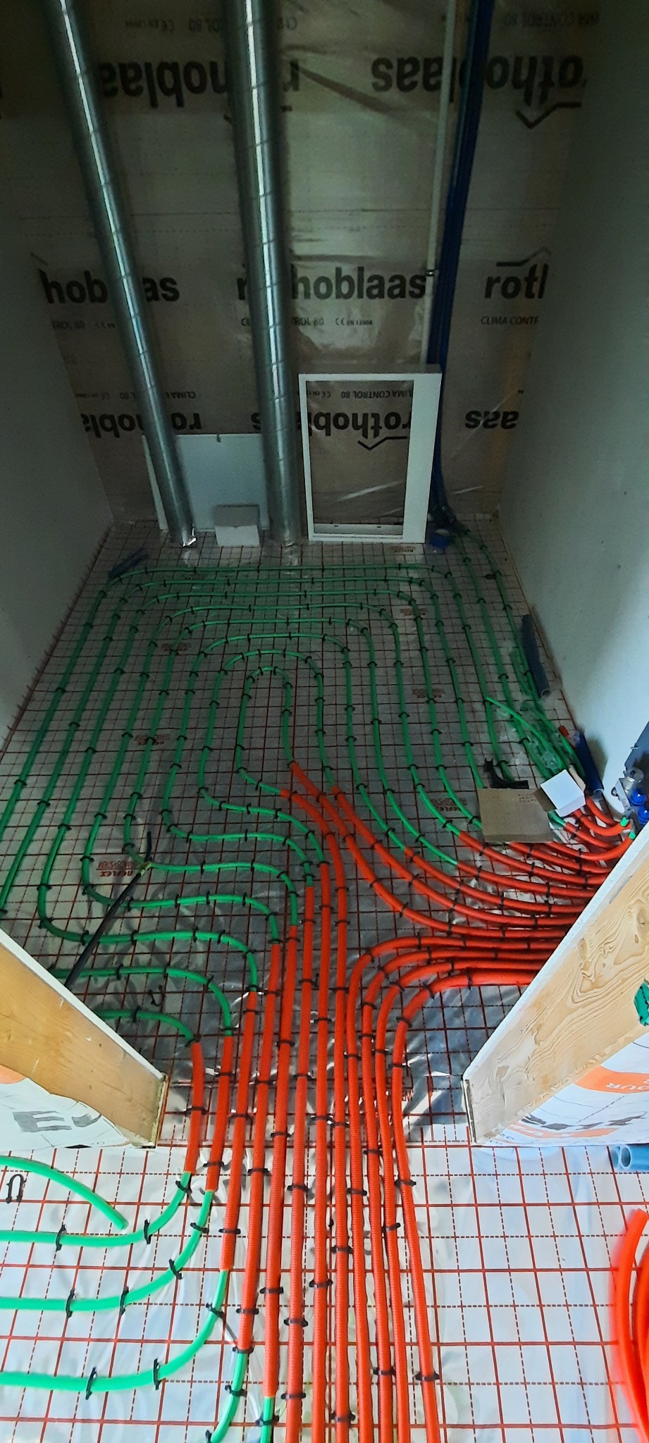 Montaż ogrzewania podłogowego: widok z góry na rozłożone rury grzewcze w kolorach zielonym i pomarańczowym, przymocowane do siatki na izolacji podłogowej. Widoczne elementy wentylacji i izolacji...